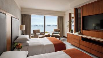 A hotel room with two beds, a TV, a desk, and a view of the sea.