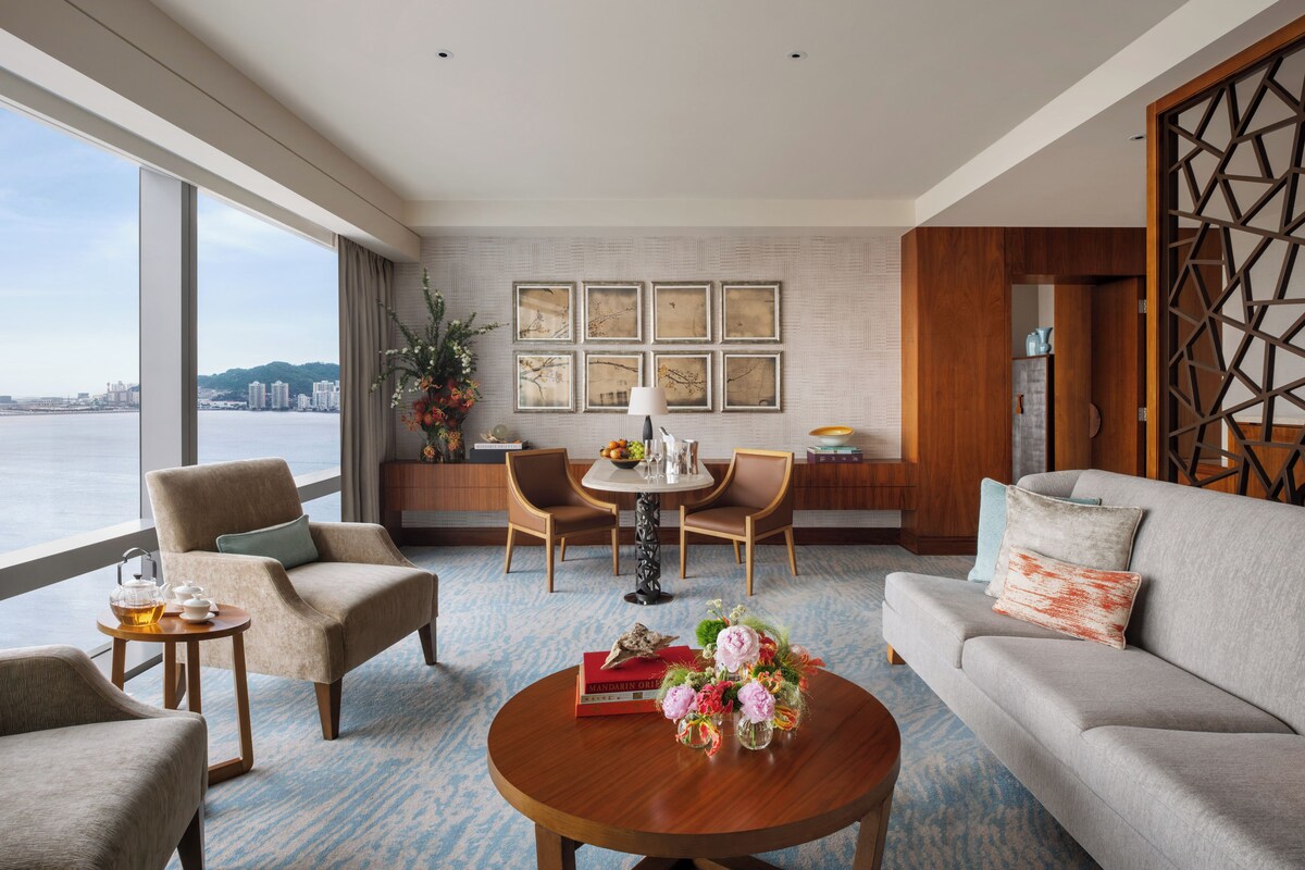 A modern hotel room with a large window offering a city view, a dining area with a table and chairs, and a seating area with a sofa and armchairs.