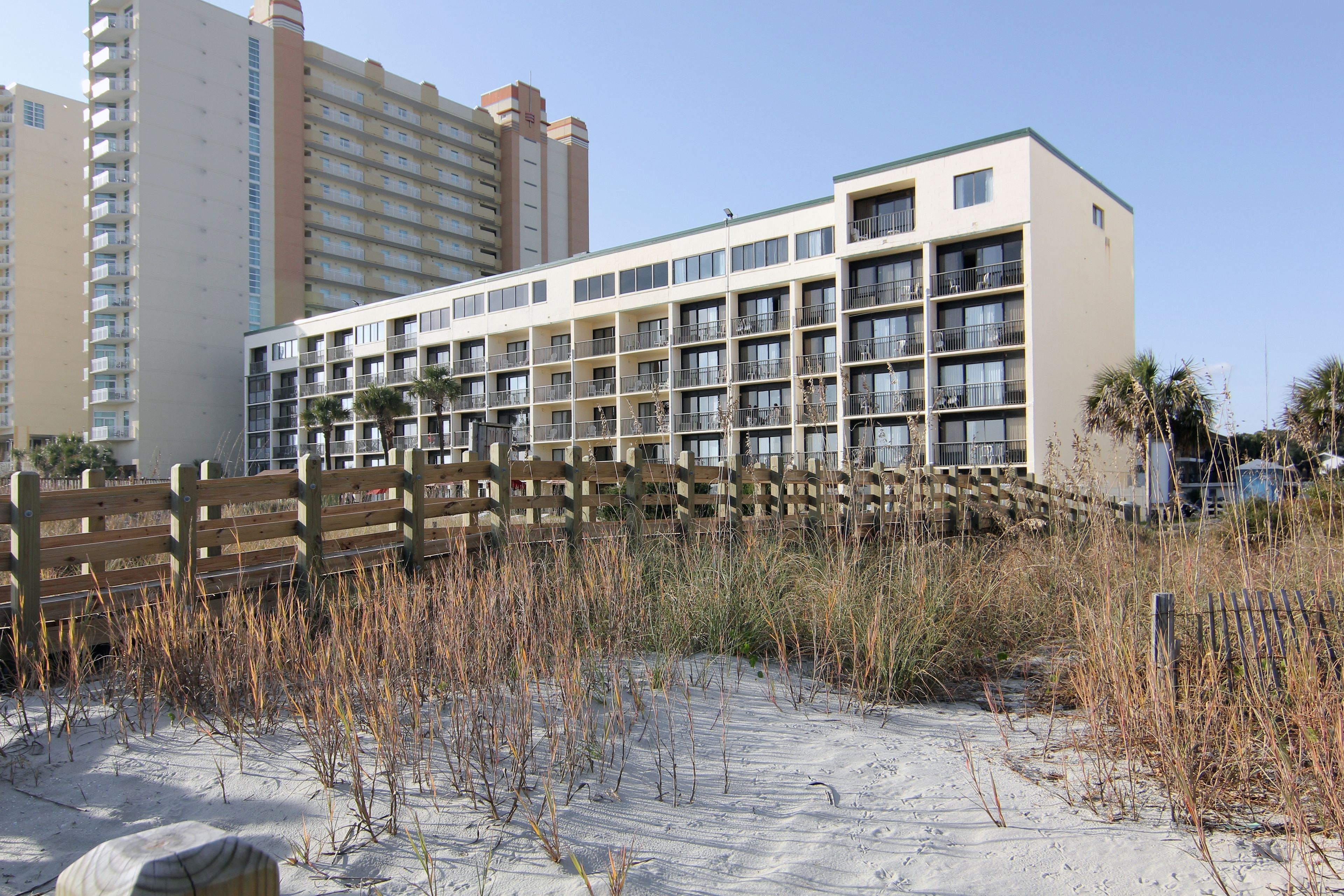 A beachfront hotel with multiple balconies, a sandy beach in front, and tall buildings in the background.