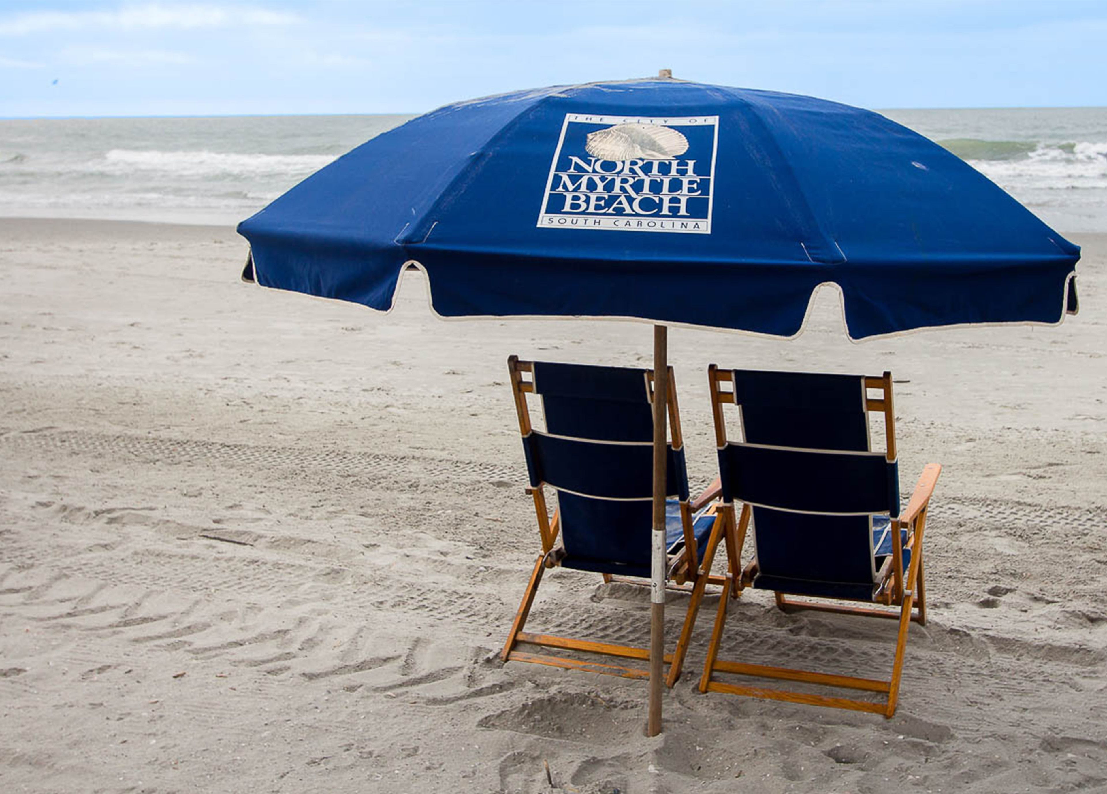 A beach umbrella with a logo, two wooden beach chairs, and a sandy beach with the ocean in the background.