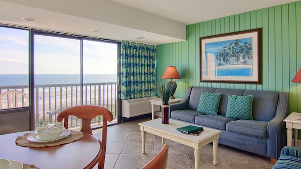 A room with a green wall, a sofa, a table, and a chair. There is a window with a view of the ocean.