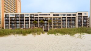 A multi-story hotel with a flat roof, balconies, and a sandy beach in the foreground.