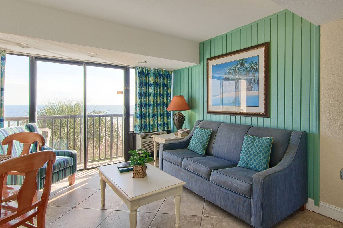 A room with a blue sofa, a wooden chair, a small table, and a view of the ocean through a sliding glass door.