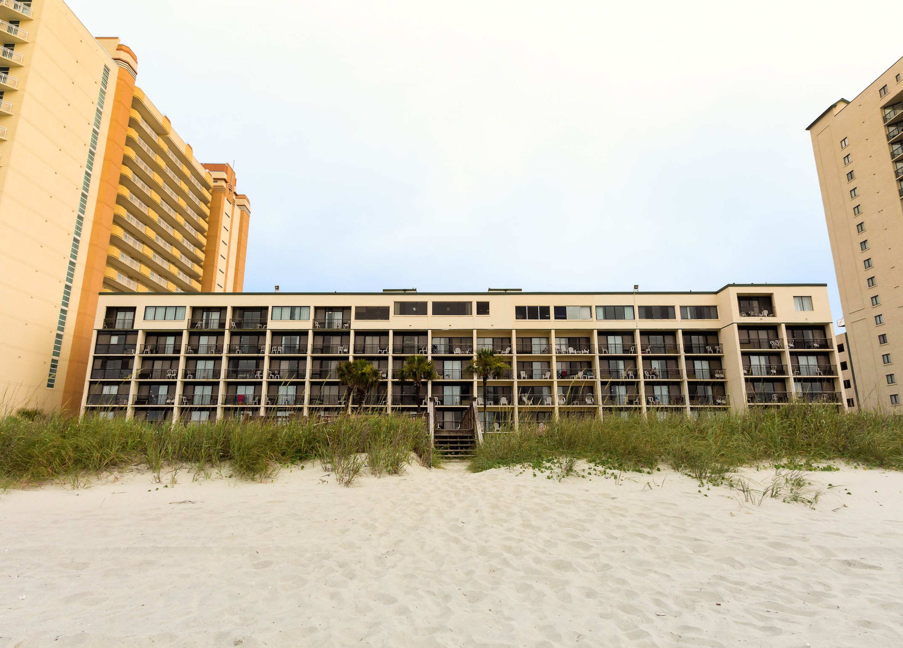 Ein mehrstöckiges Hotel mit schlichter Fassade, vielen Balkonen und einem Sandstrand im Vordergrund.