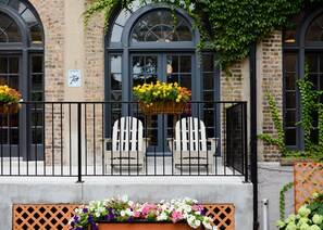 Un bâtiment aux murs de briques, doté de grandes fenêtres en arc de cercle et d’un balcon agrémenté de deux fauteuils Adirondack blancs.