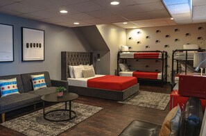 Una habitación de hotel moderna con un sofá gris, una cama con ropa de cama roja, una mesita con una planta y una litera con ropa de cama roja.