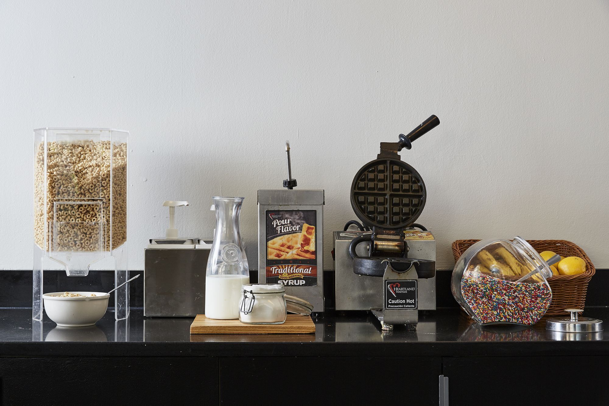 A kitchen counter with a waffle maker, a bottle of syrup, a jar of sugar, a bowl of cereal, and a basket of fruit.