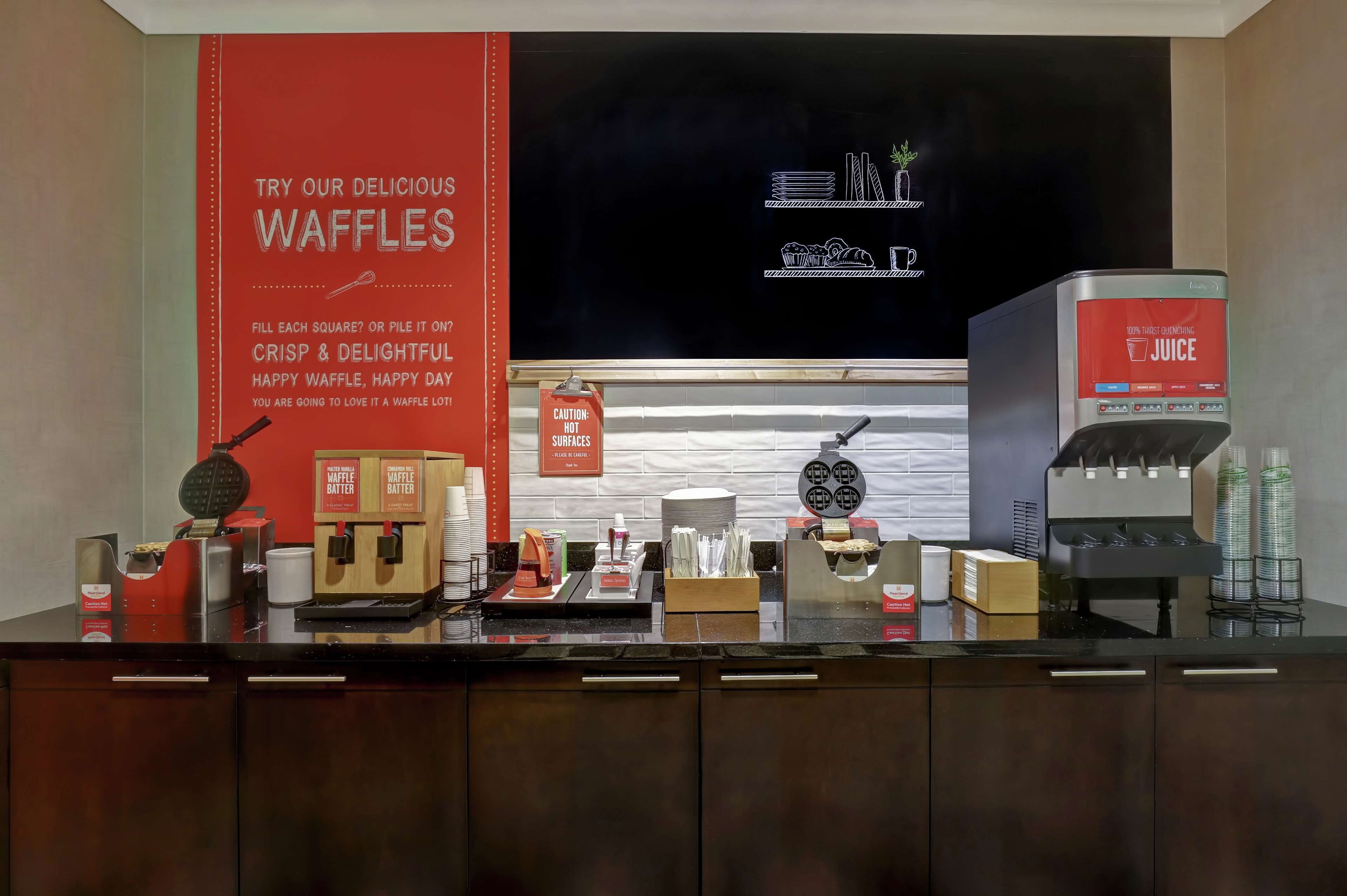 Un buffet à gaufres avec un panneau rouge, des boîtes de pâte à gaufres, un appareil à gaufres et un distributeur de jus.