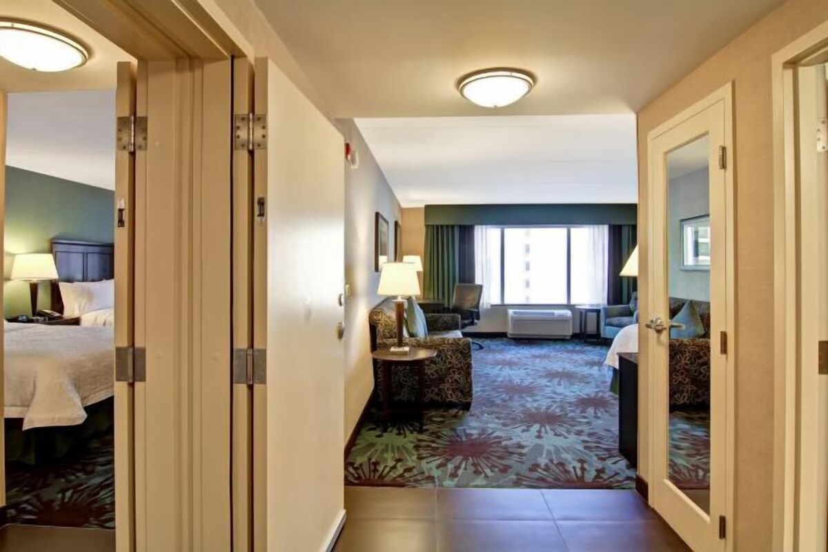 A hotel room with a bed, a sofa, a small table with a lamp, and a large window.