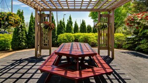 Una mesa de picnic techada con una pérgola encima, rodeada de abundante follaje y cestas de flores colgantes.