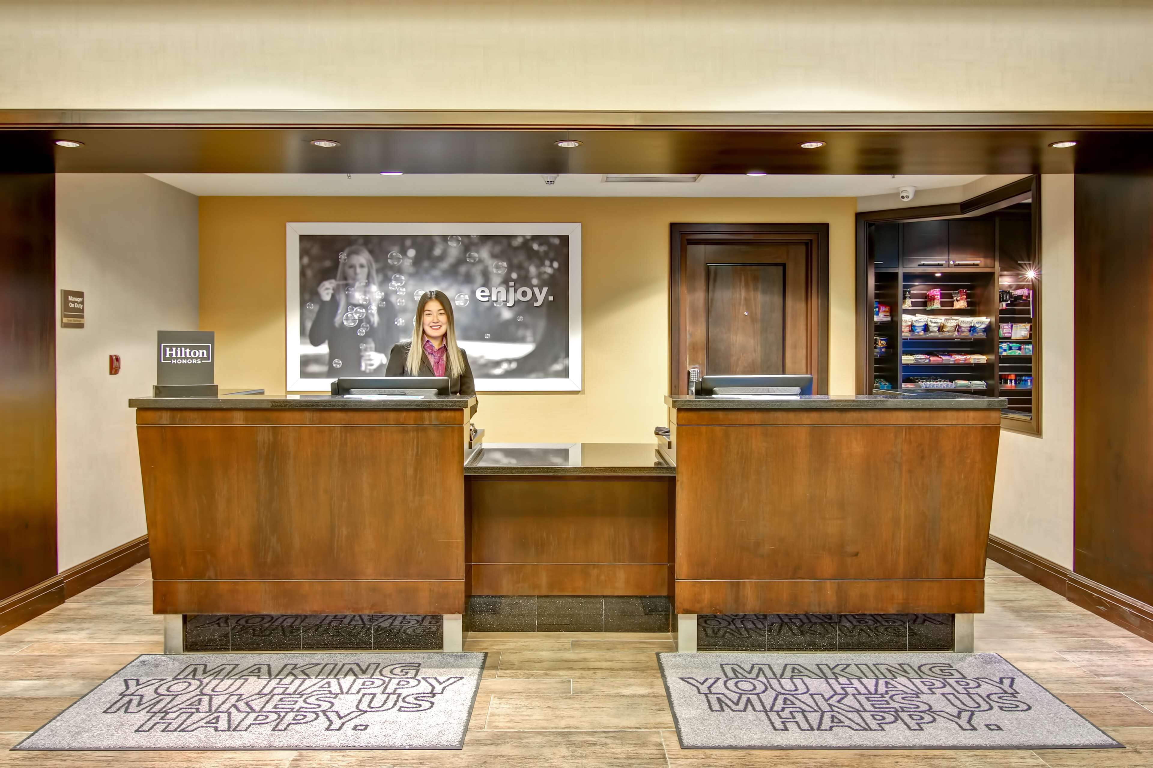A hotel reception area with two wooden reception desks, a large 'enjoy.' poster, and a 'Making you happy makes us happy.' mat.