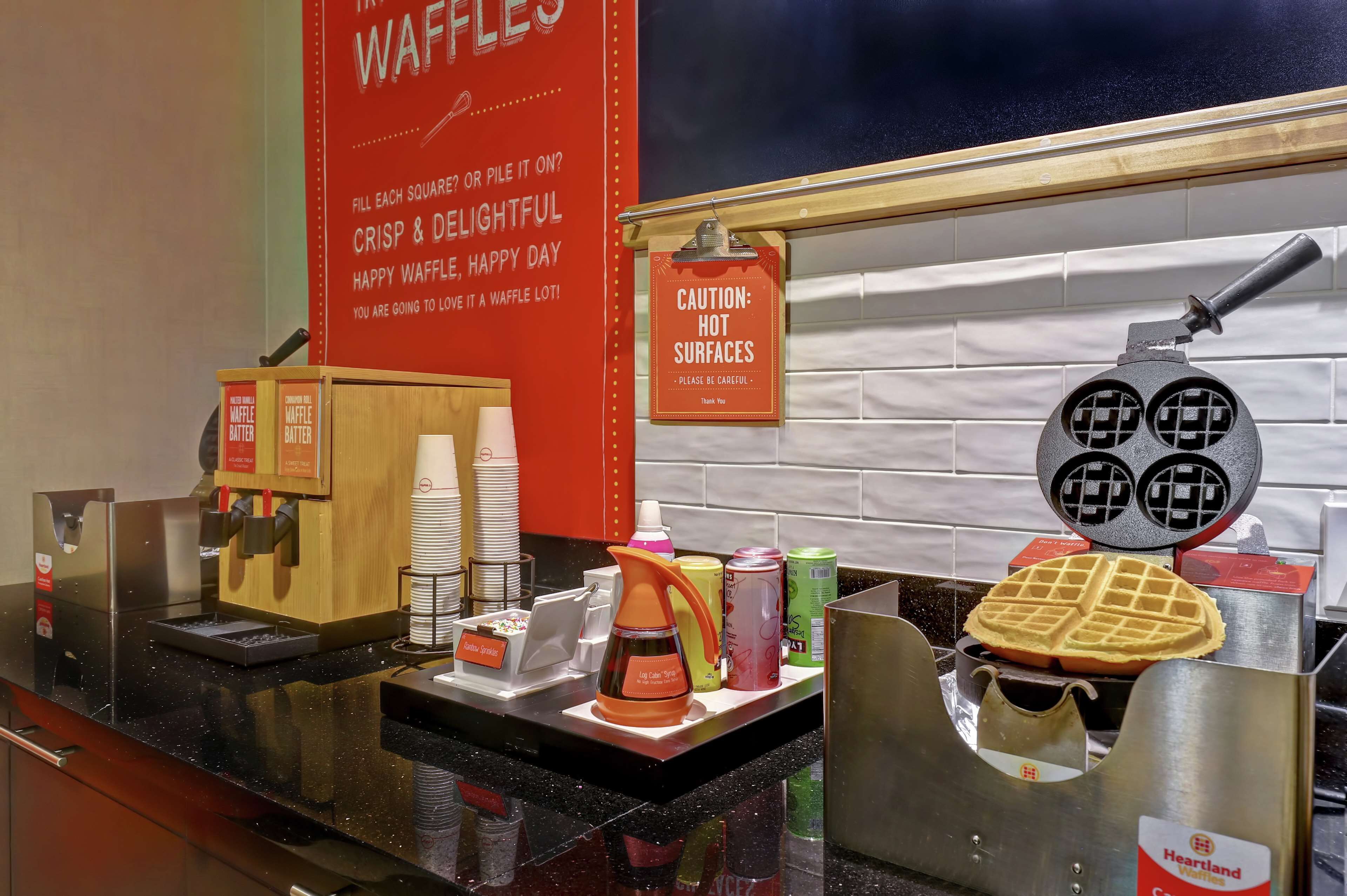 Una estación de gofres con una máquina para hacer gofres, dispensadores de masa para gofres y una señal de precaución por superficies calientes.