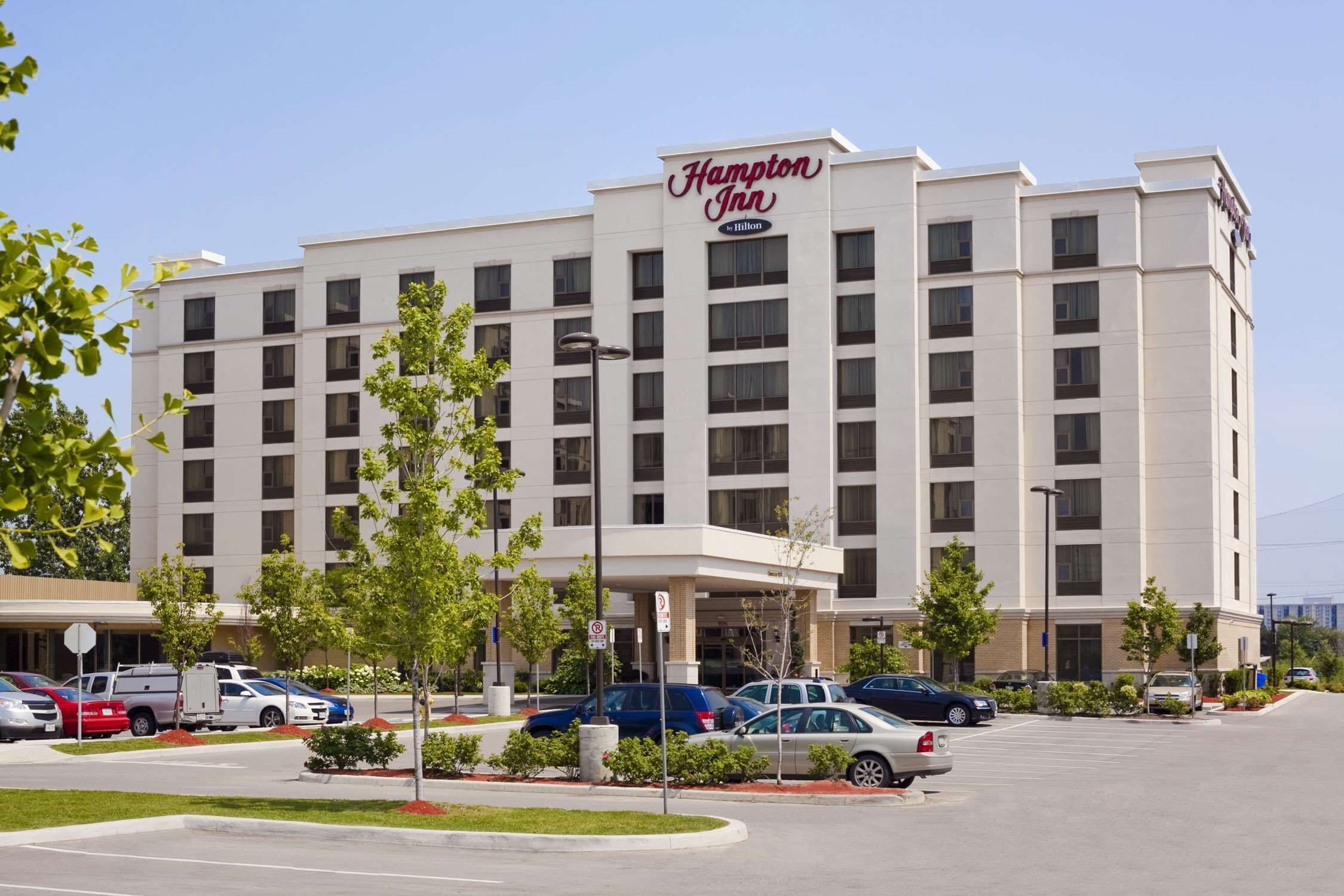 A Hampton Inn hotel with a modern design, featuring a prominent entrance and a parking lot with several cars.