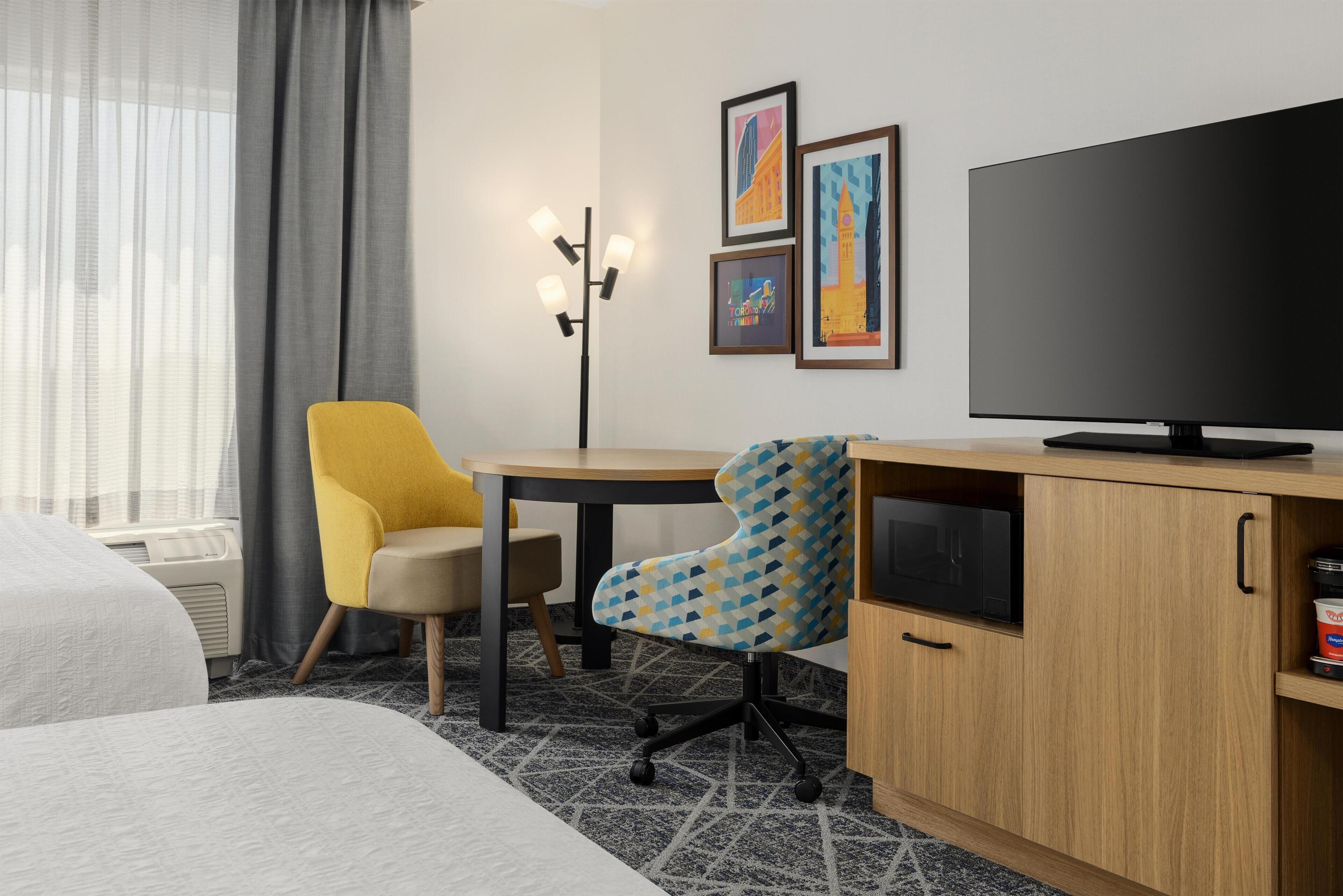 Habitación de hotel con cama, escritorio con computadora, silla, televisor y obras de arte en la pared.