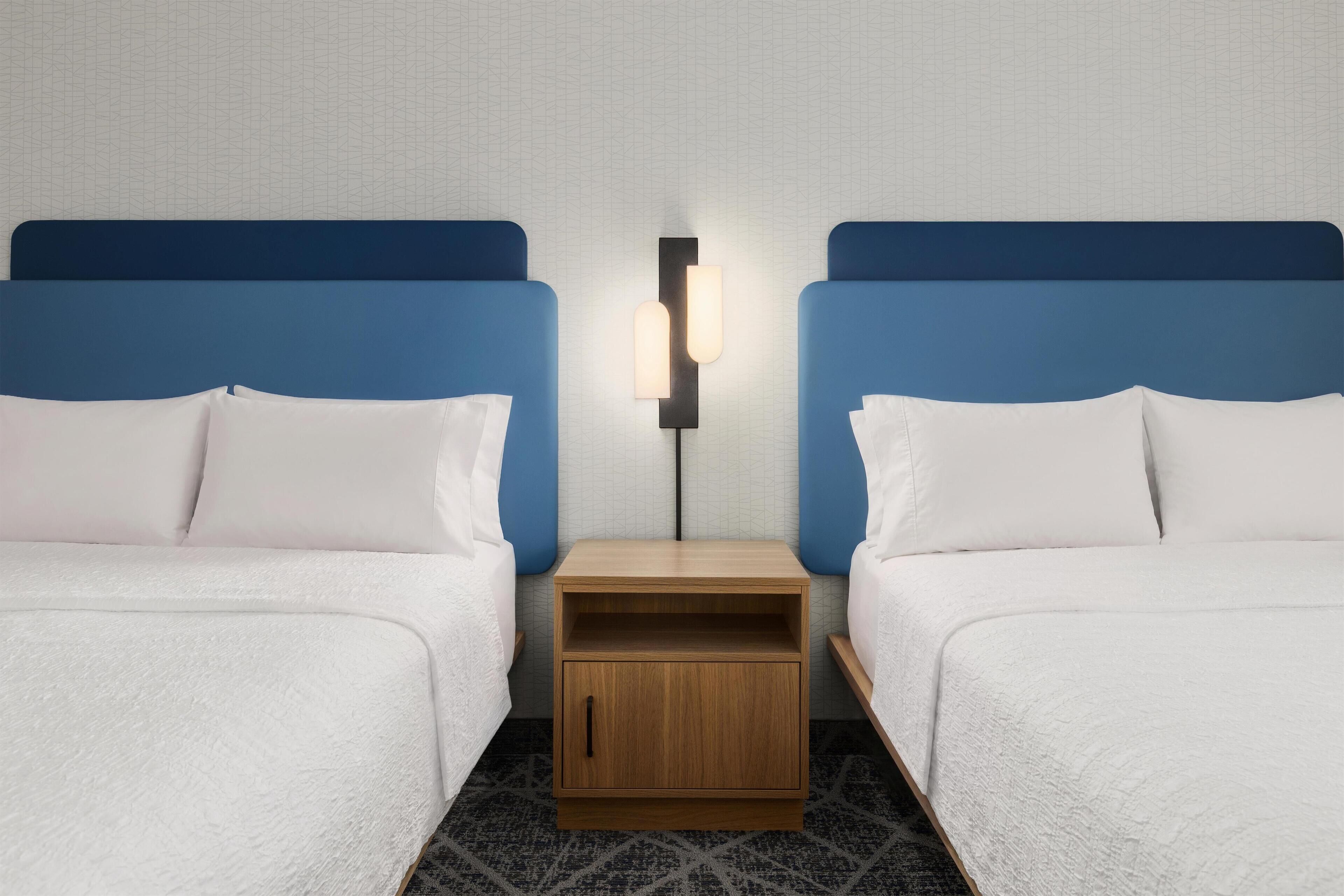 A hotel room with two beds, a nightstand, and a wall-mounted light fixture.