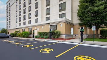 Un estacionamiento con estaciones de carga para vehículos eléctricos, un edificio con múltiples ventanas y un árbol.