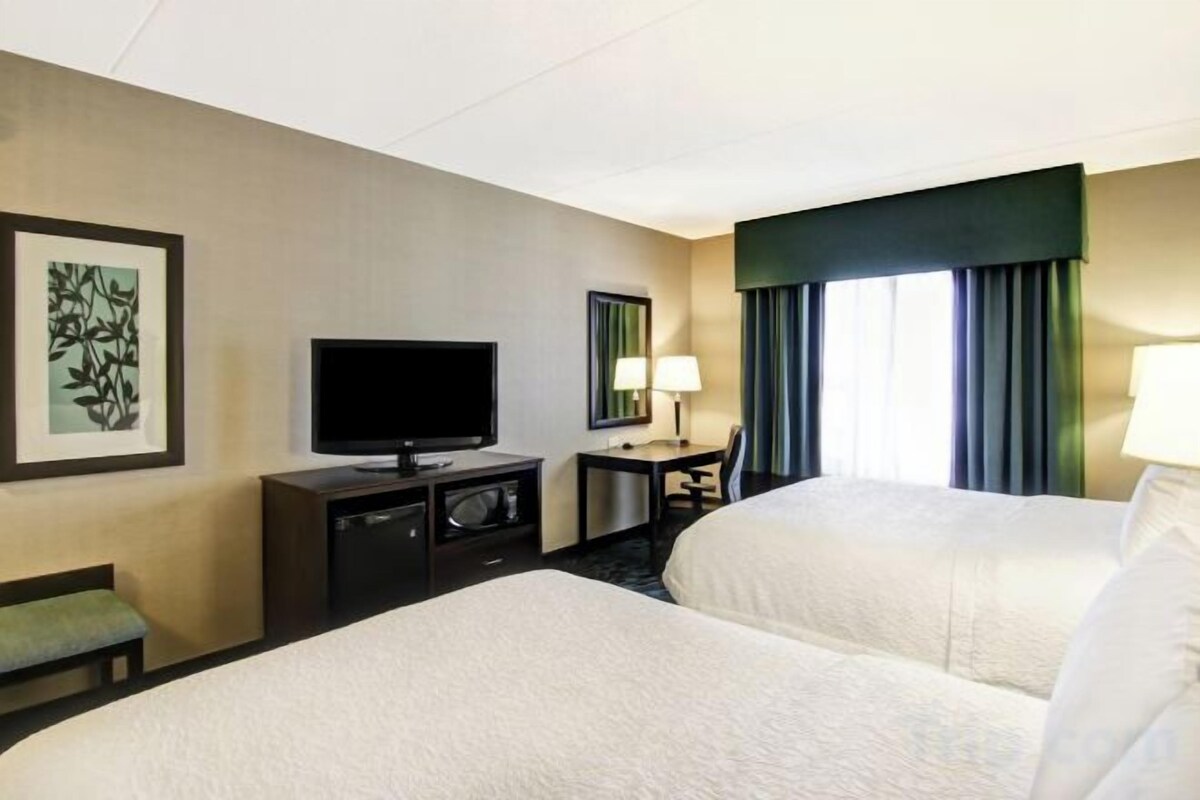 Habitación de hotel con dos camas, un televisor de pantalla plana, un escritorio con lámpara y una ventana con cortinas.