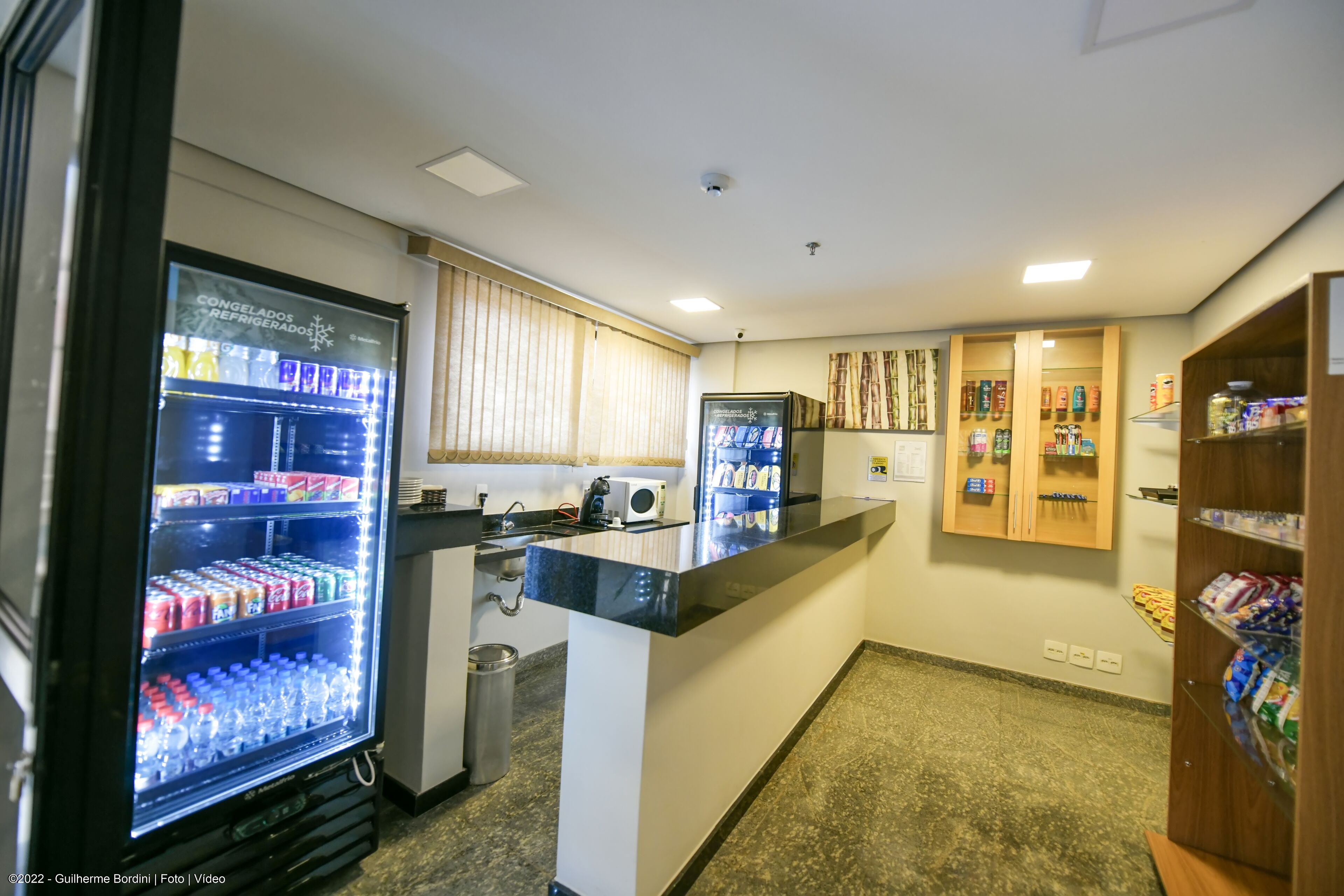 A hotel lobby with a reception desk, two refrigerators stocked with beverages, a trash bin, and a wooden shelf with snacks.
