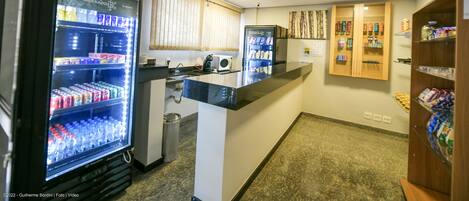 A hotel lobby with a reception desk, two refrigerators stocked with beverages, a trash bin, and a wooden shelf with snacks.