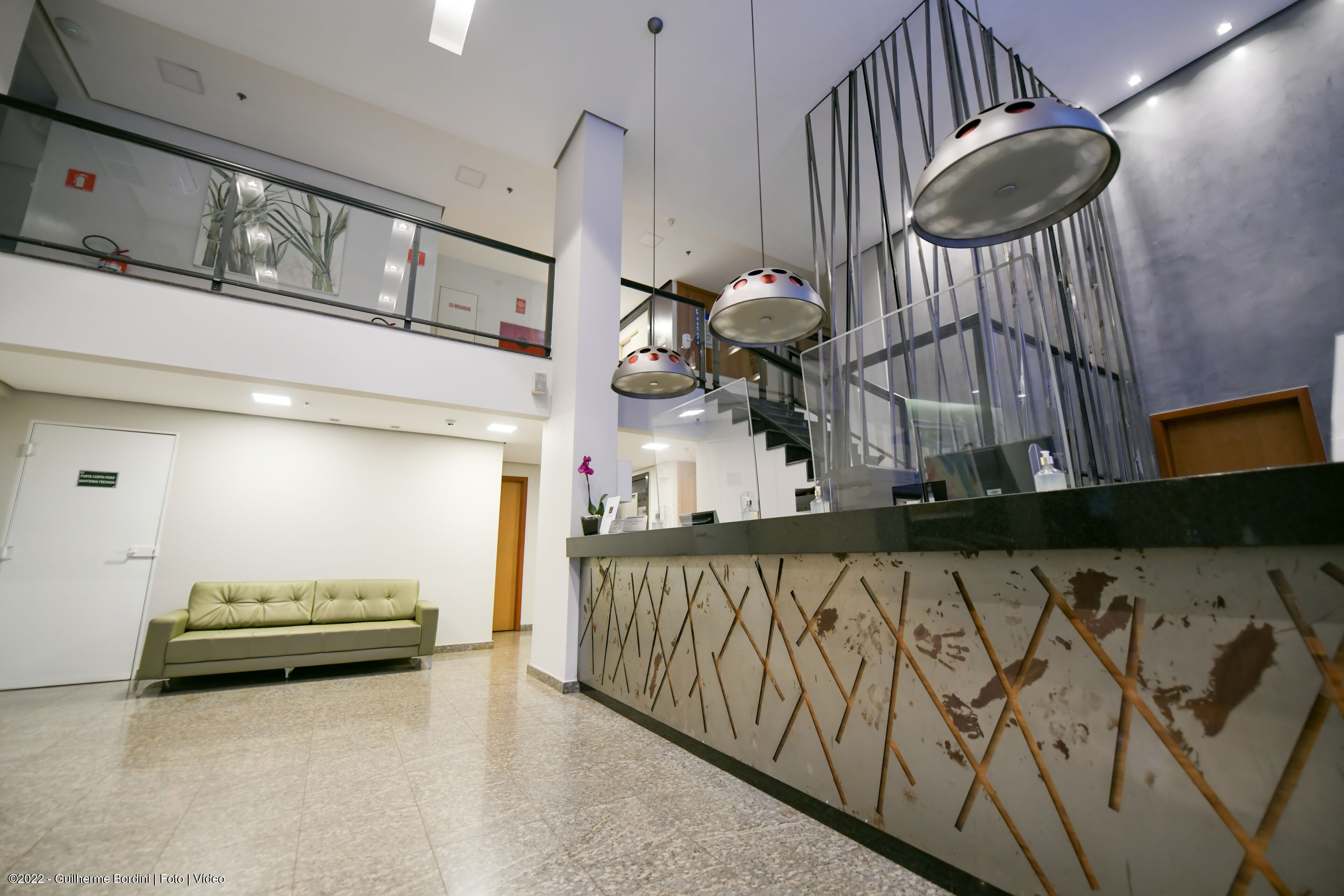 Un hall con mostrador de recepción, un sofá verde, una escalera e iluminación colgante moderna.
