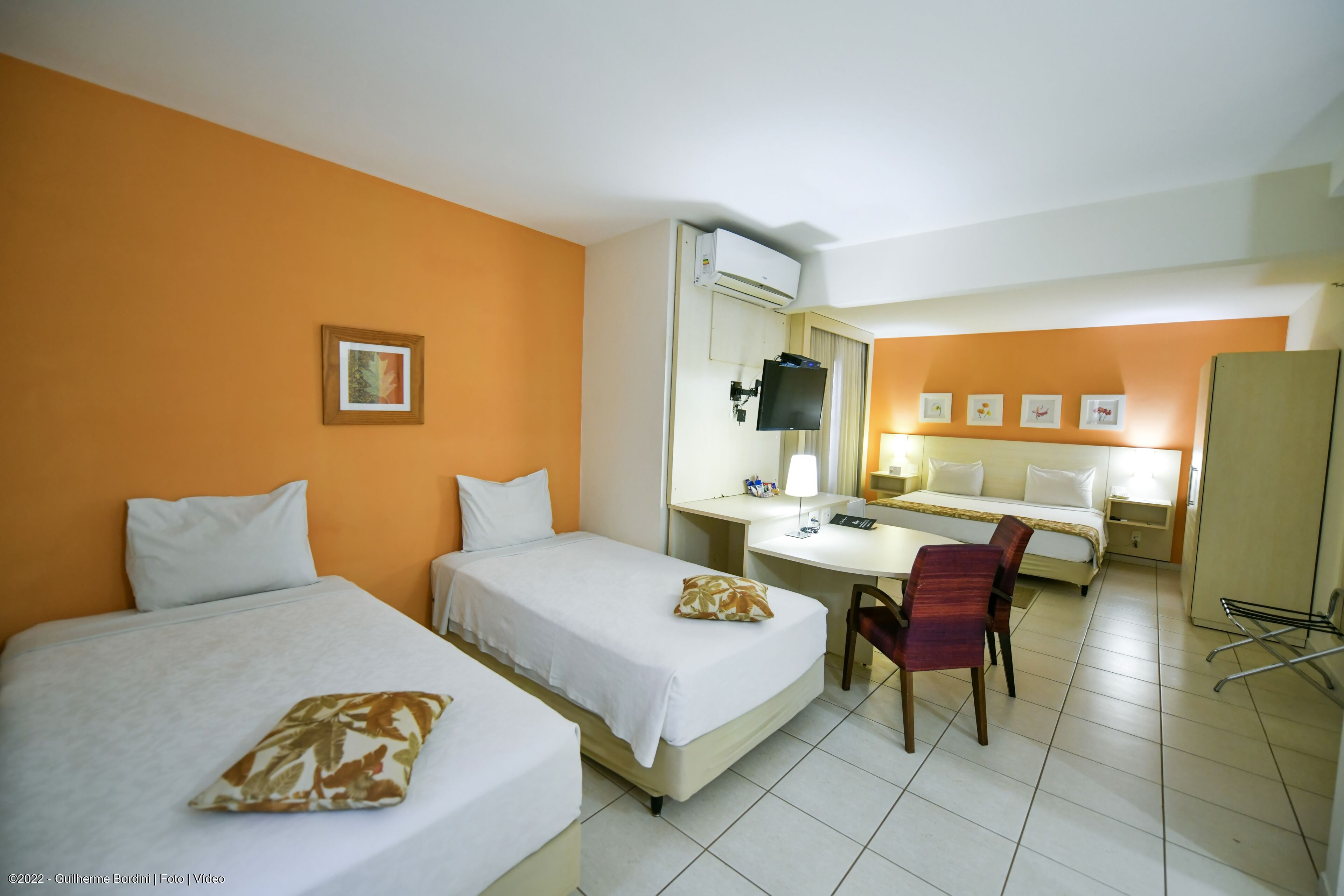 Quarto de hotel com duas camas, uma escrivaninha, uma cadeira, uma televisão e um guarda-roupa.