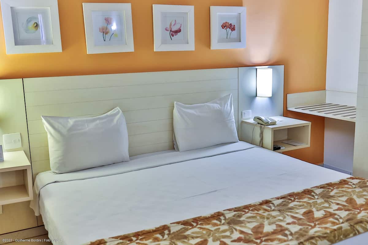 A hotel room with a bed, white pillows, a bedside table with a phone, and framed floral artwork on the wall.