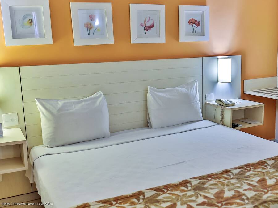 Ein Hotelzimmer mit einem Bett, weißen Kissen, einem Nachttisch mit Telefon und gerahmten floralen Bildern an der Wand.