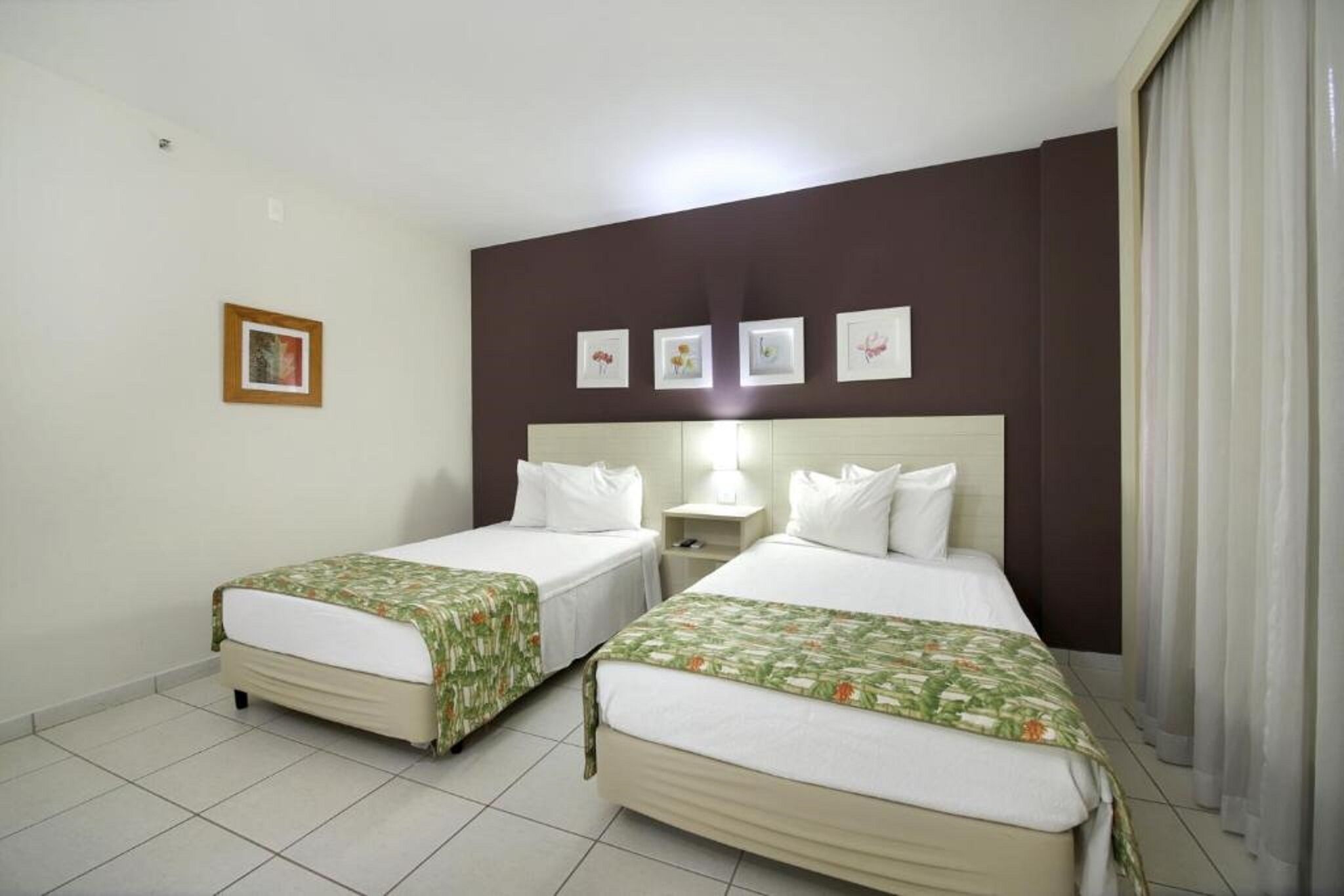 A hotel room with two single beds, a nightstand, a wall-mounted shelf, and a window with curtains.