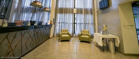 A hotel reception area with a reception desk, two armchairs, a small table with a lamp, and a television on the wall.