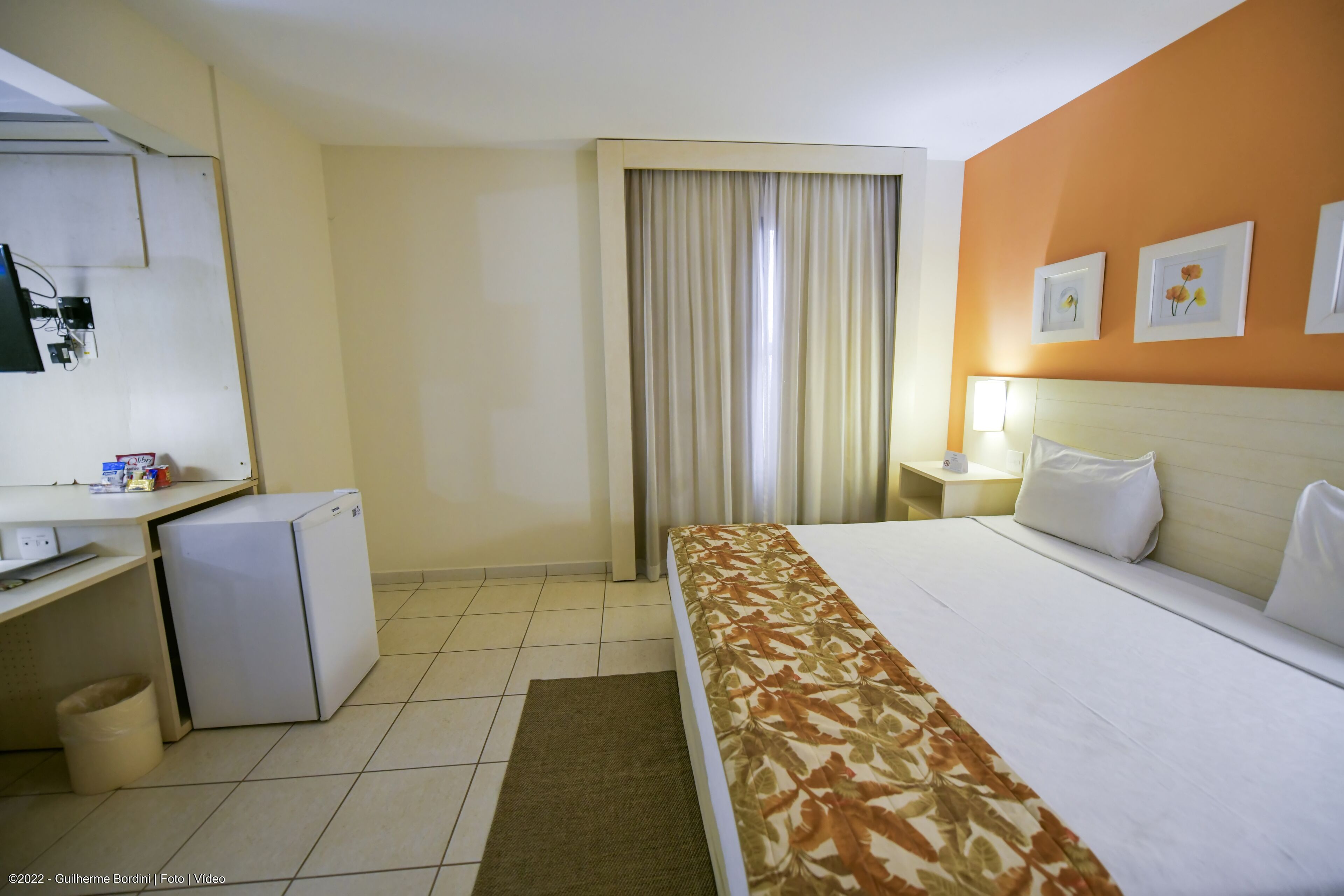 A hotel room with a bed, a desk with a computer, a small refrigerator, a window with curtains, and a nightstand with a lamp.