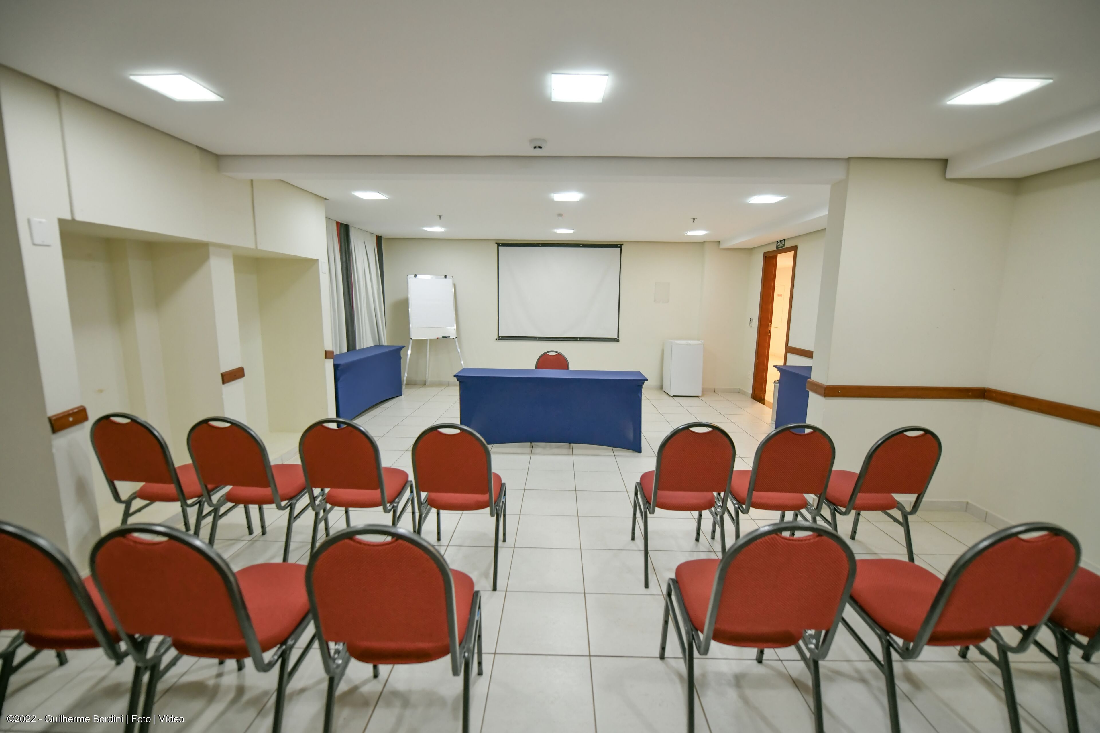 A conference room with a blue table, red chairs, a whiteboard, and a projector screen.