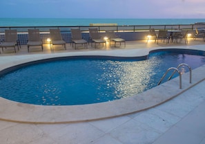 A swimming pool with lounge chairs and tables, a tiled deck, and a view of the ocean.