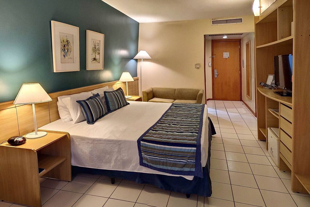 A hotel room with a bed, two bedside lamps, a desk with a computer, a TV, and a wardrobe.