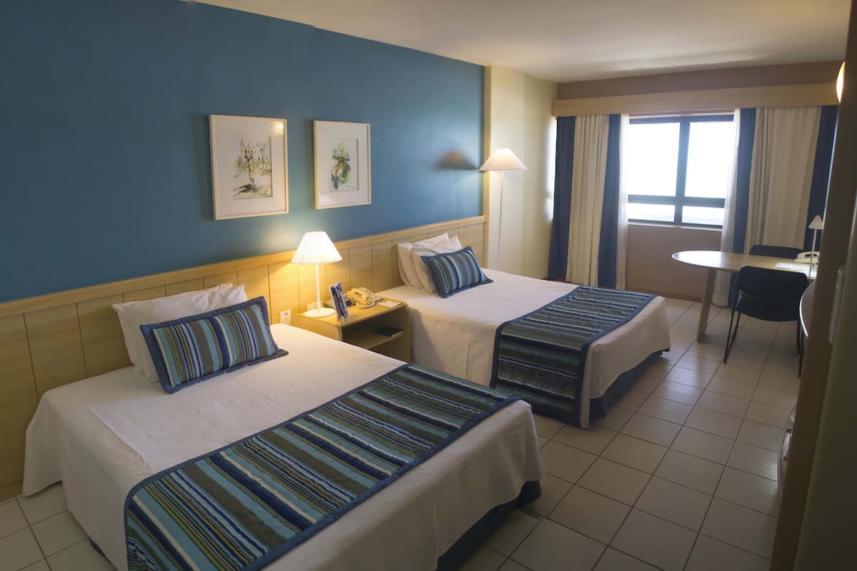 A hotel room with two beds, a small table, and a chair. There are two framed pictures on the wall, a lamp on each nightstand, and a window with curtains.
