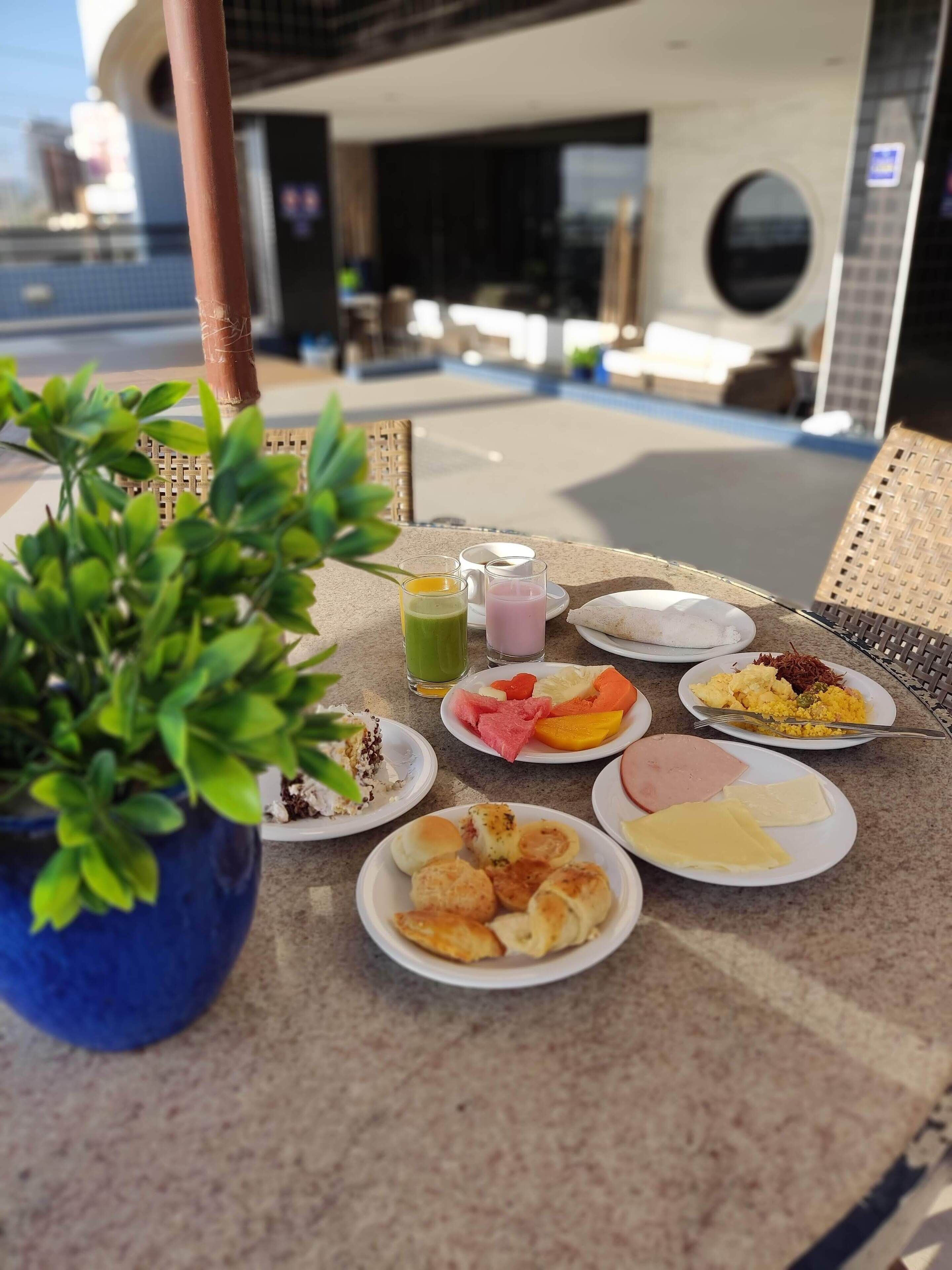Mesa servida con diversos productos para el desayuno, como frutas, pasteles y bebidas, con vista a una piscina y un edificio al fondo.