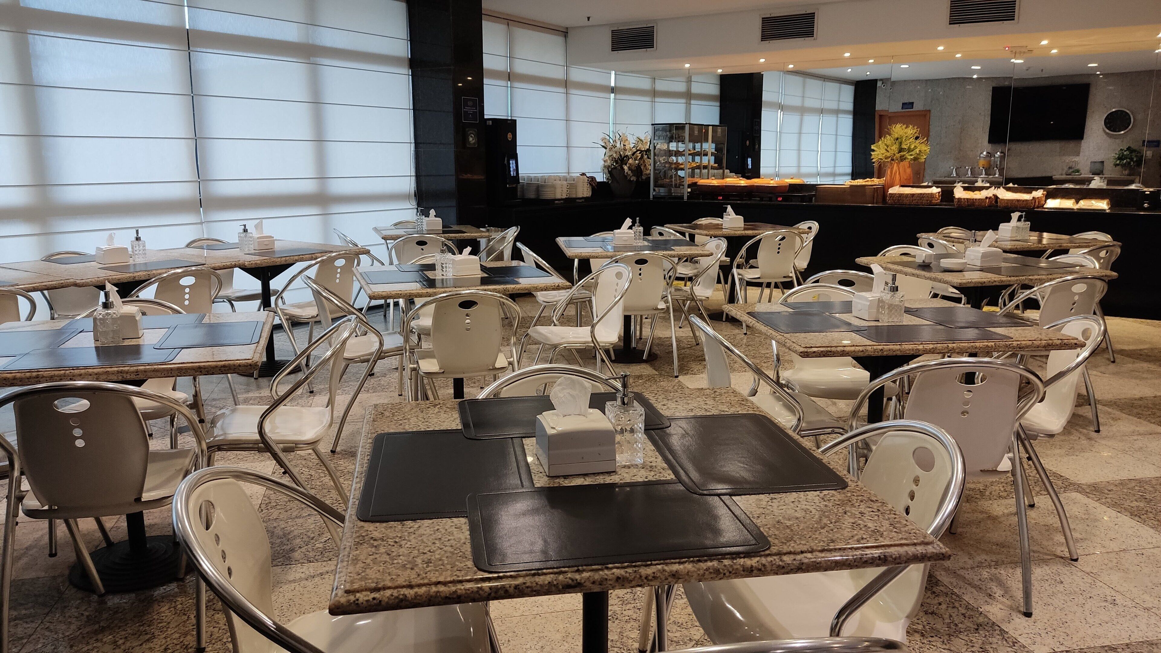 Un comedor de hotel con varias mesas y sillas, una zona de bar y amplias ventanas que permiten la entrada de luz natural.