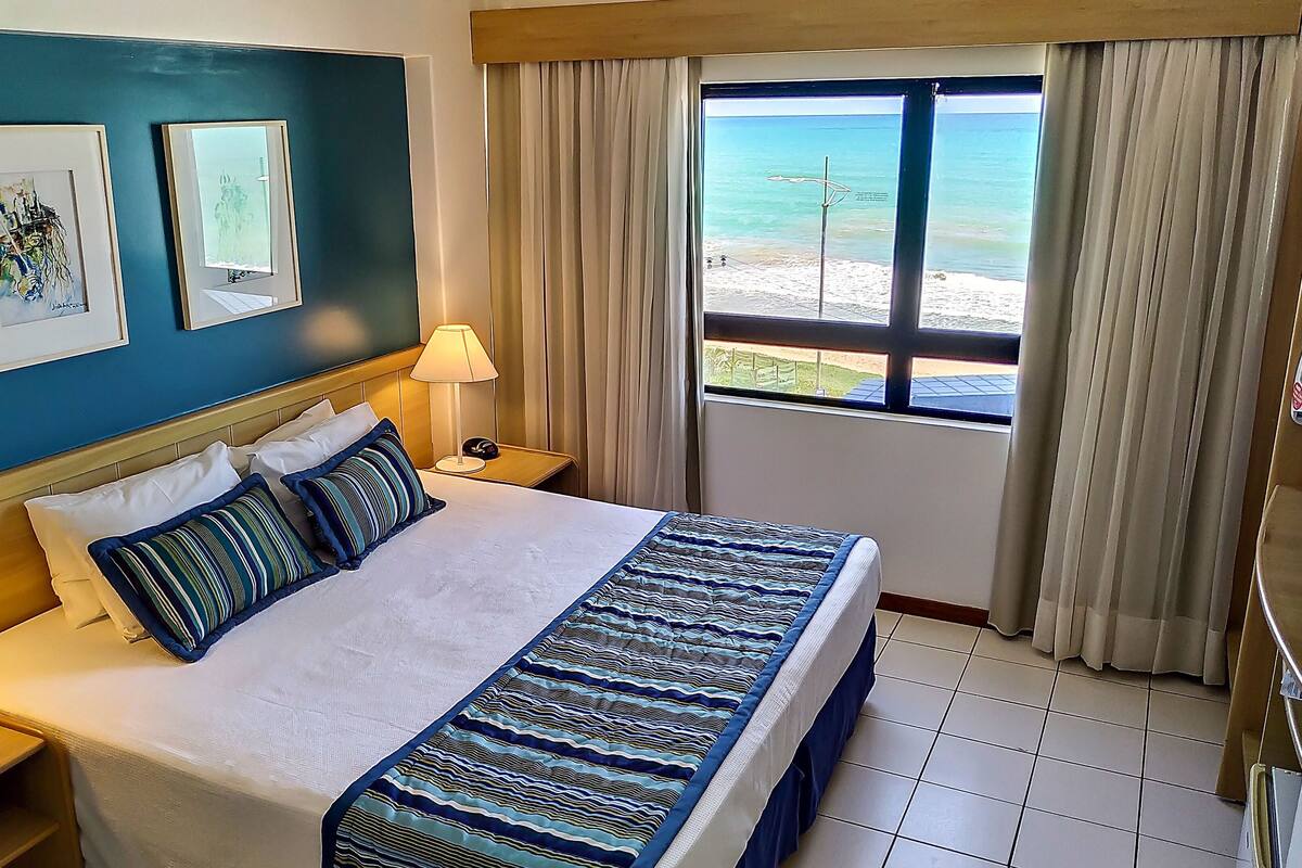 A hotel room with a bed, two framed artworks, a window with a beach view, a lamp, and a nightstand.