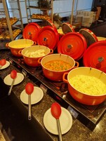 Montaje de bufé con diversos platos en ollas rojas y amarillas, junto con cucharones rojos sobre platos blancos.