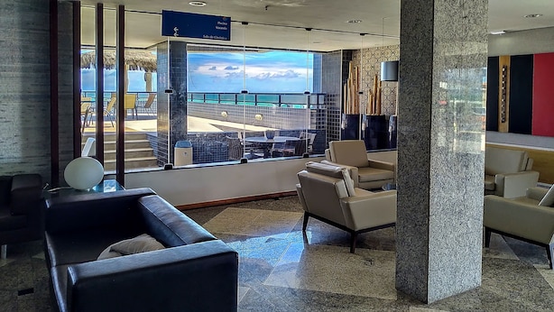 A modern lobby with a large window offering a view of a pool and outdoor seating. There are several chairs and a small table. - Reymar Hotel Maceió (Maceió)