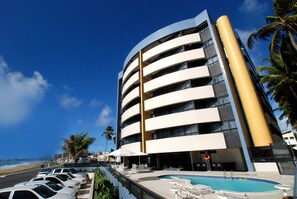 A modern hotel with a curved facade, a pool area with lounge chairs, and a beach view.