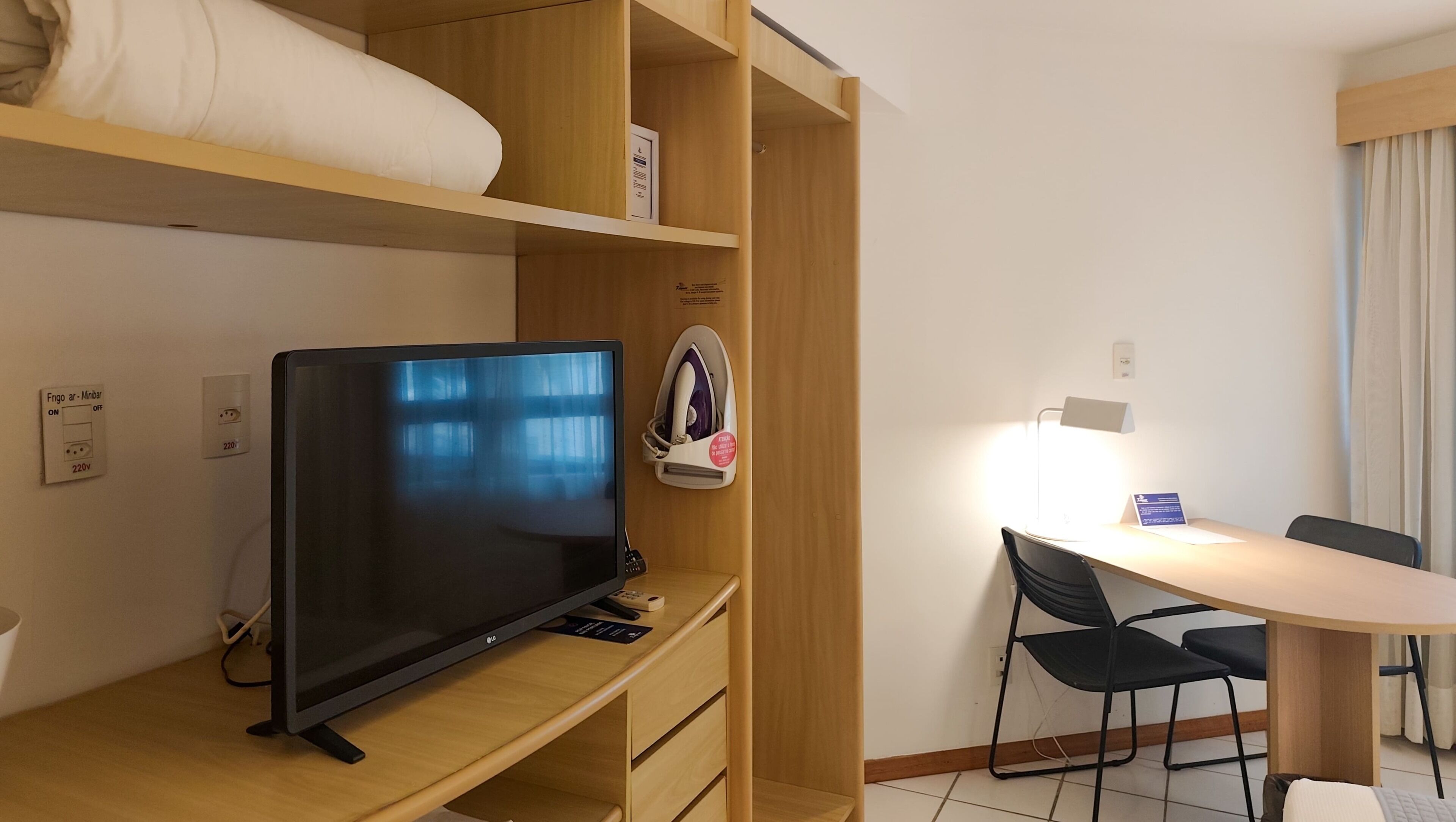 A hotel room with a desk, a television, a chair, a bed, and a small table with a lamp.