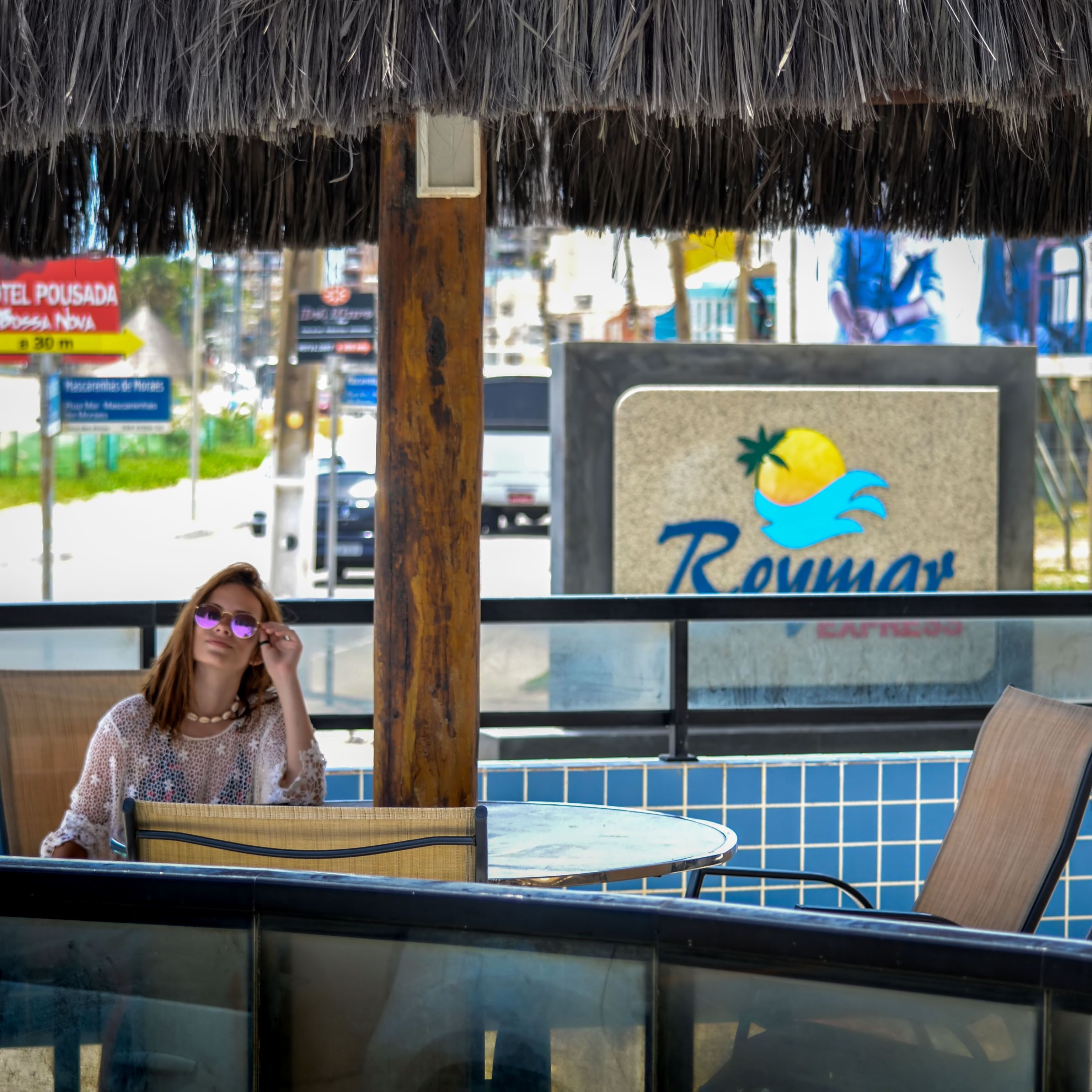 A person sitting at a table with a thatched roof overhead. There is a sign with the word "Rovem" and a palm tree logo.