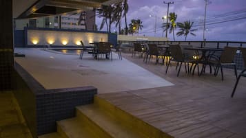 Terraço no telhado com piscina, área de estar e vista para palmeiras e um edifício ao fundo.