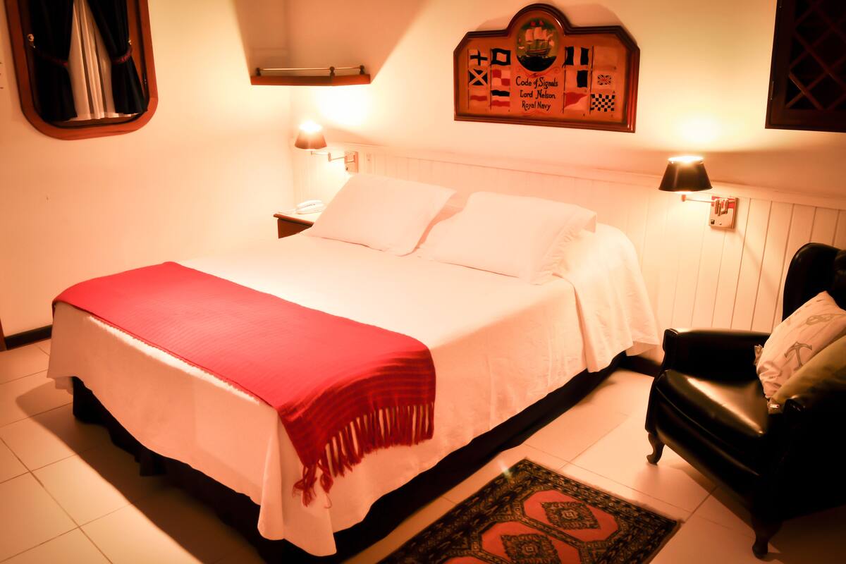 Una cama bien hecha con una colcha roja, una butaca de cuero negro y un reloj fijado a la pared.