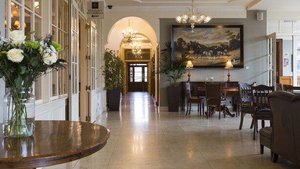 A hotel lobby with a long corridor, a chandelier, a dining area with wooden furniture, and a large painting on the wall.