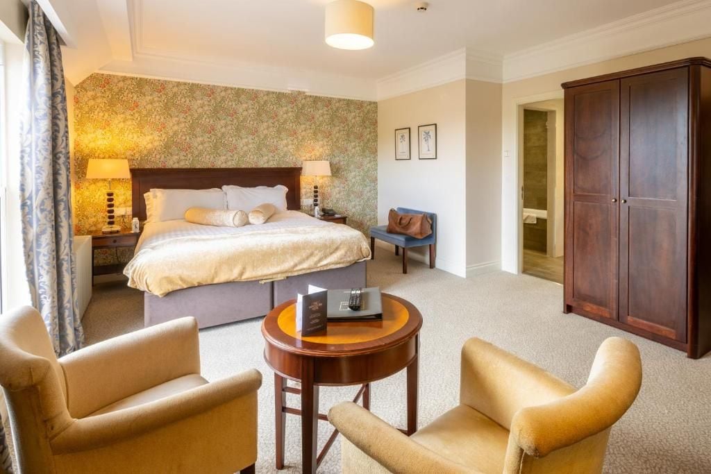 A hotel room with a bed, bedside tables, a desk, two armchairs, a wardrobe, and a carpeted floor.