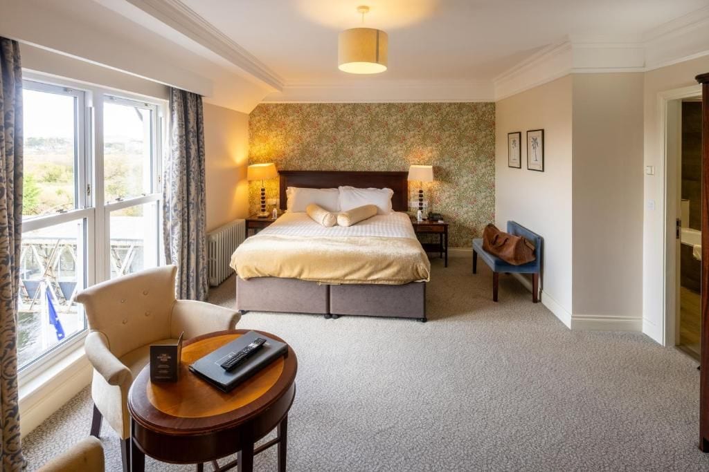 A hotel room with a large bed, a sitting area with a table and chair, a view through a window, and a door leading to another room.