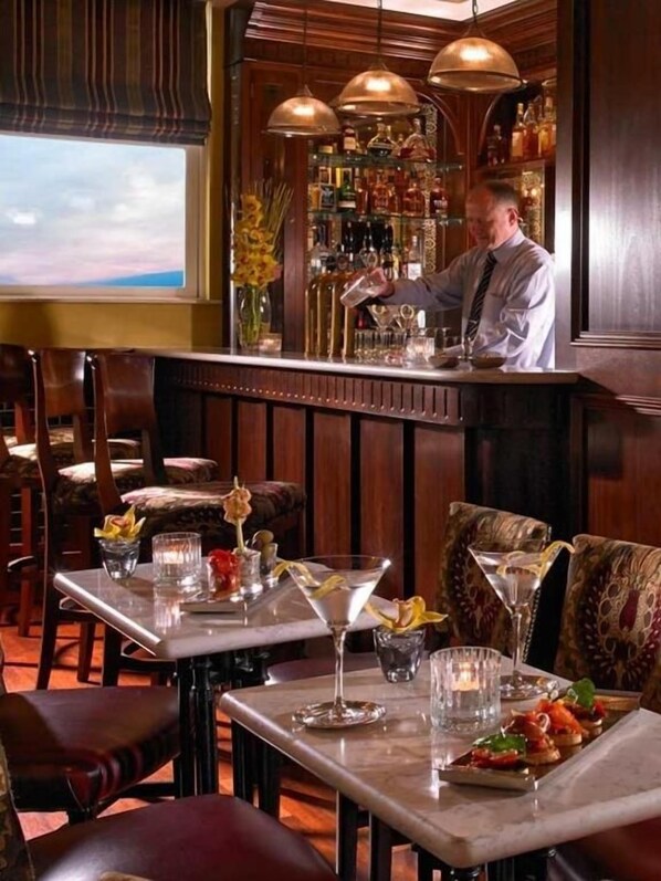 A bar area with a bartender, bar stools, and a bar counter. There are tables with chairs and a view of the sky through the window.