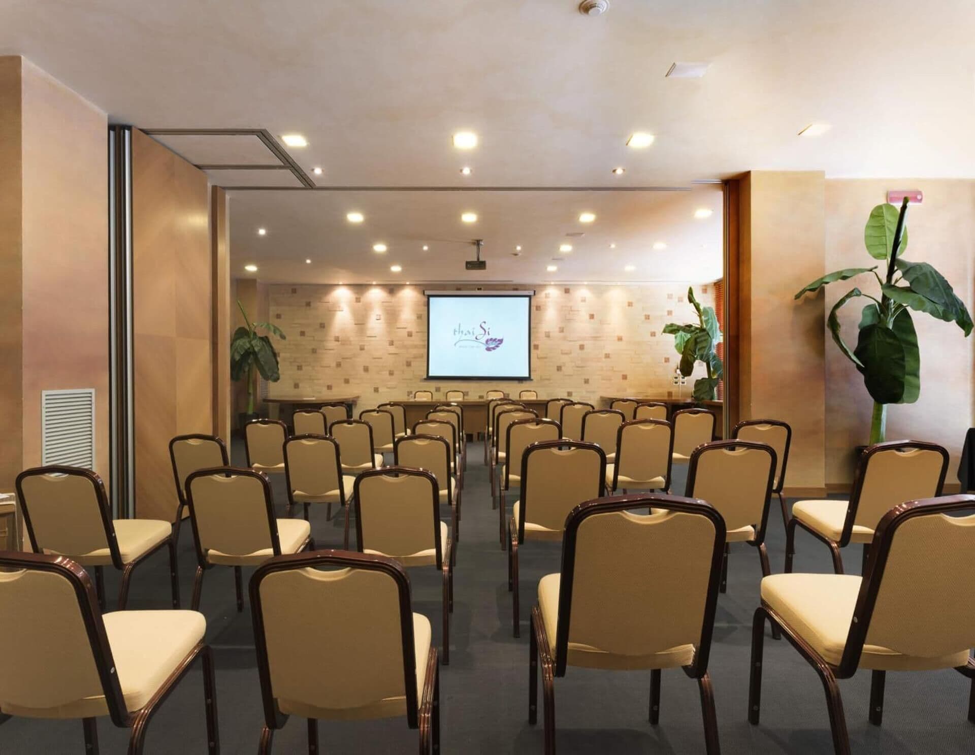 Une salle de conférence avec des rangées de chaises disposées face à un écran de projection sur lequel est affiché le titre « Thai Si ».