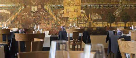 A restaurant with tables set for dining, a mural depicting traditional architecture, and a view of a temple.